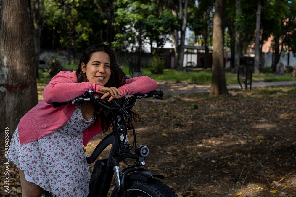 Obraz premium Young Latin American woman (33) goes for a walk in spring in the park with her electric bicycle, the sun illuminates her beautiful face. Concept means of transport and spring.