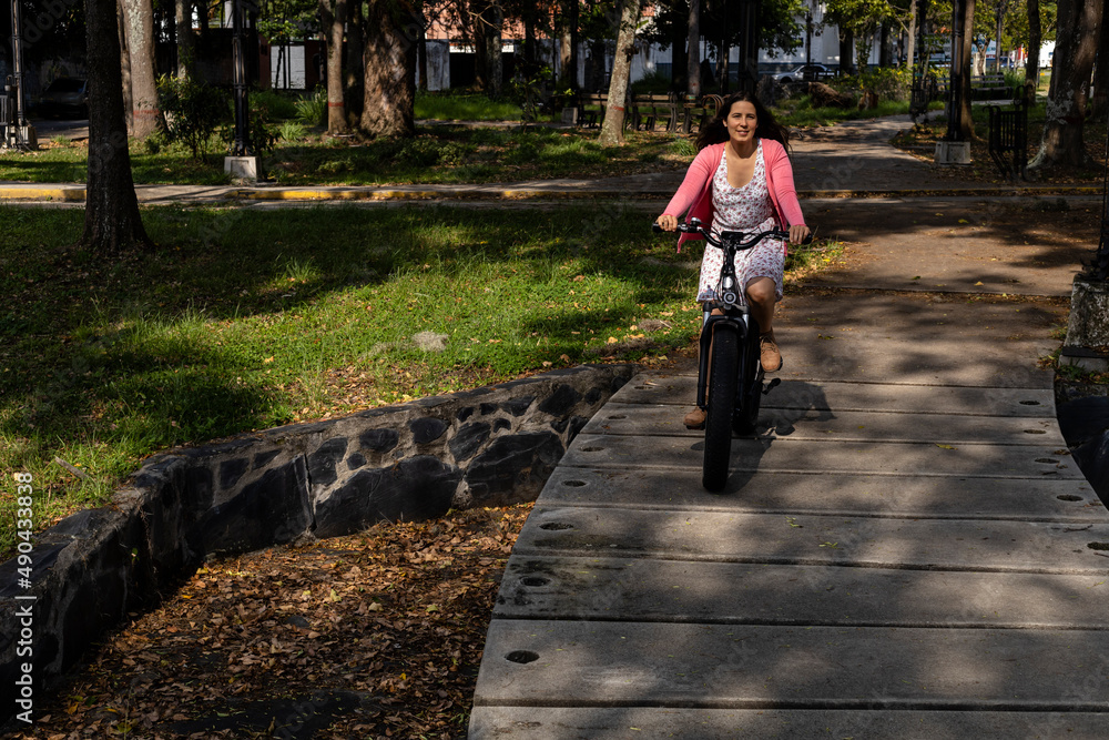 Obraz premium Young woman (33) Latin American is riding her electric bicycle in the park, spring sun illuminates her face. Concept means of transport and spring.