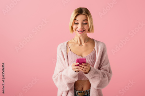 happy young woman in soft cardigan using smartphone isolated on pink.