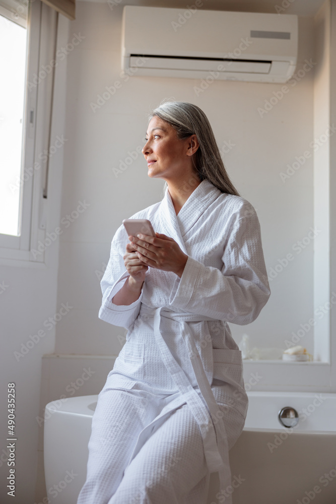 Pretty Asian woman in bathrobe holding smartphone in bathroom Pretty Asian woman in bathrobe holding smartphone in bathroom