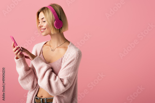 cheerful woman listening music in wireless headphones and using smartphone isolated on pink.