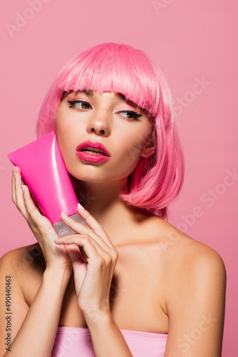 young woman with colored hair holding tube with lotion isolated on pink.