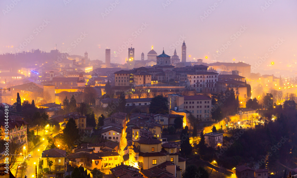 Picturesque night view of old fortified Upper City of Bergamo on hill with brightly lit streets and historic buildings in winter haze, Italy