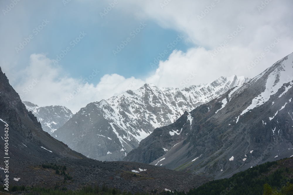 Awesome landscape with high snowy mountain range with sharp rocks in cloudy sky. Dramatic view to snow mountains in changeable weather. Atmospheric mountain scenery with white snow on black rocks.