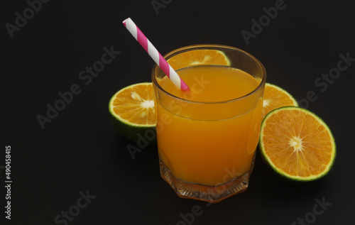 Glass of fresh orange juice with drink straw and green orange fruits slices on black background, copy space for text.