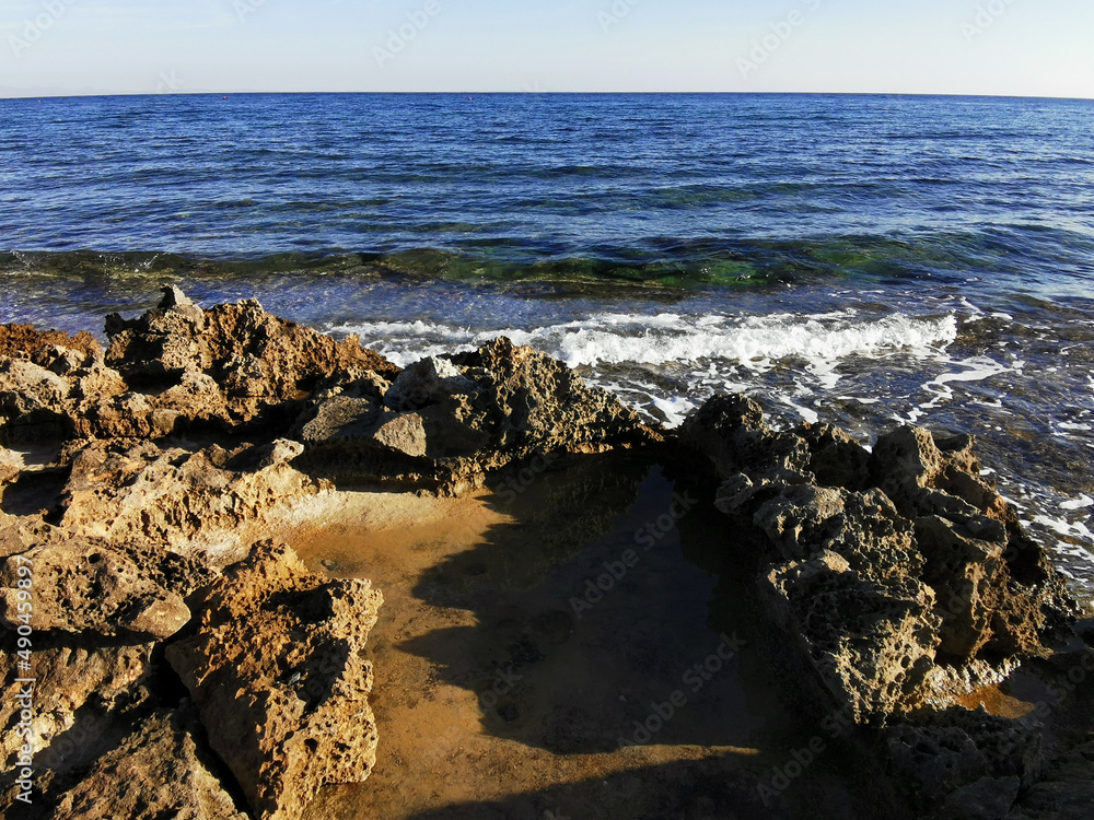 Long-hardened lava, porous, sharp coast of the Mediterranean Sea with oncoming waves against a skys.