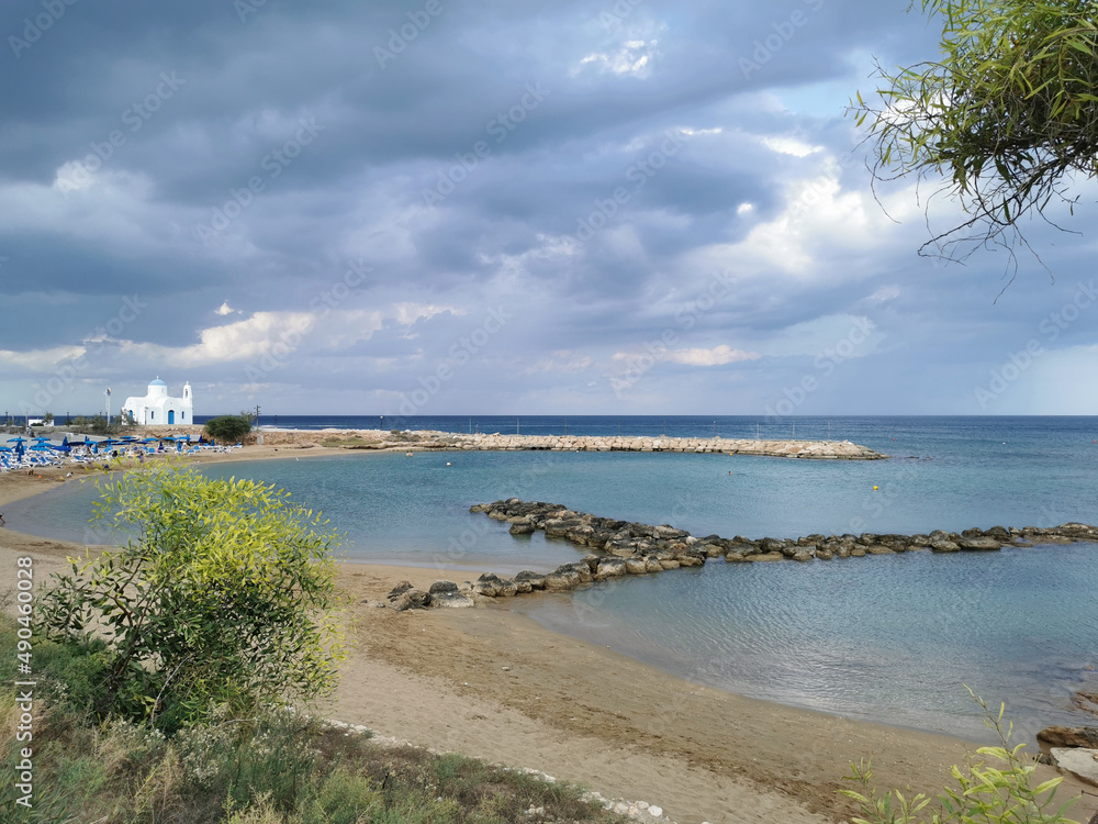 Sandy shore with a stone ridge, Kalamies beach, the Church of St. Nicholas the Wonderworker against the backdrop of a dramatic sky.