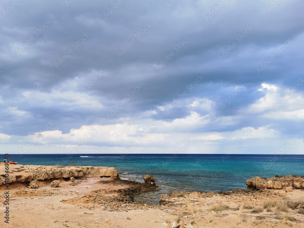 The sandy coast of the Mediterranean Sea, turning into stones from long-hardened lava, azure water with a white boat against a dramatic sky.