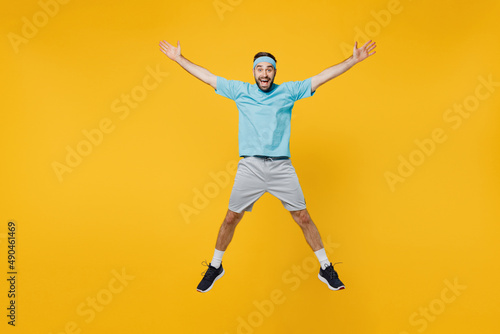 Full body young fitness trainer instructor sporty man sportsman in headband blue t-shirt jump high with outstretched hands legs isolated on plain yellow background. Workout sport motivation concept.