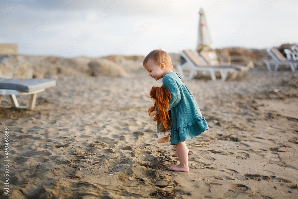 Cute european baby toddler in a green dress plays with a doll on the beach by the sea at sunset, gray background. Girl hugging waldorf soft doll, favorite toys and summer vacation kids Cute european baby toddler in a green dress plays with a doll on the beach by the sea at sunset, gray background. Girl hugging waldorf soft doll, favorite toys and summer vacation kids