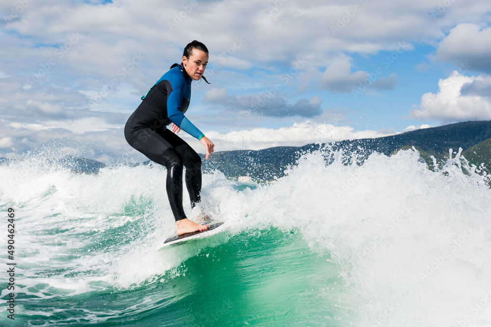 Young athletic female in wetsuit riding on endless waves behind a boat on sunny day. Woman practice wakeboarding, carving behind the baot. Watersport concept. Young athletic female in wetsuit riding on endless waves behind a boat on sunny day. Woman practice wakeboarding, carving behind the baot. Watersport concept.
