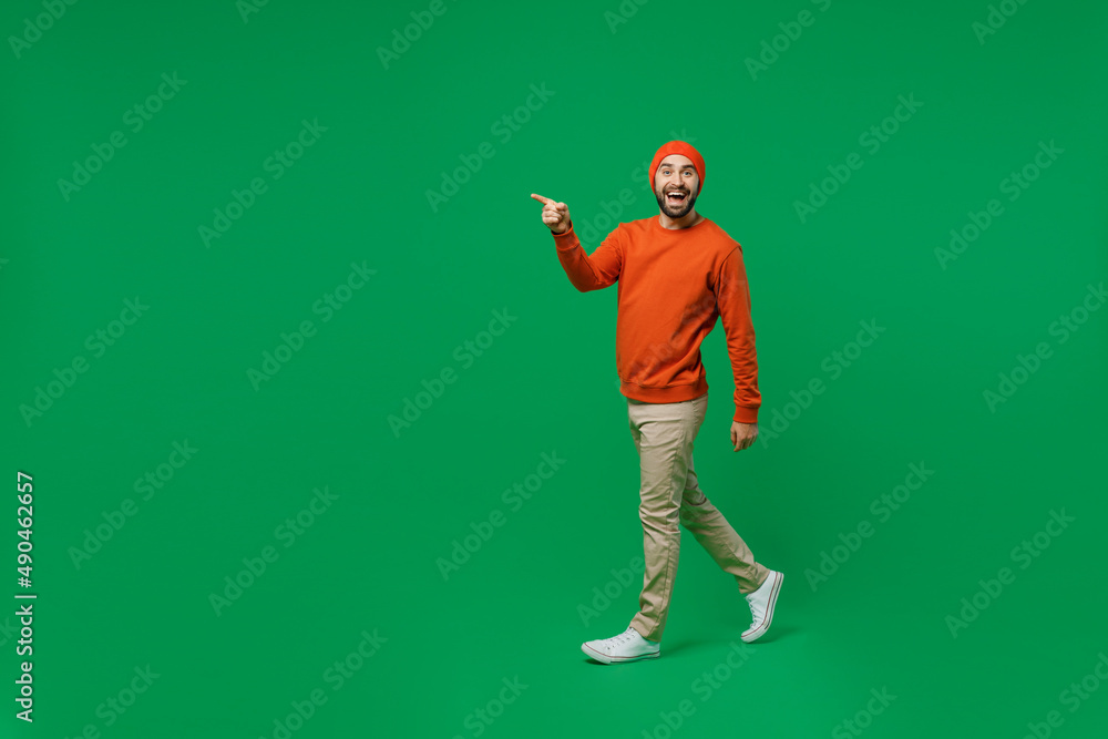 Full body young smiling happy cheerful surprised man 20s wear orange sweatshirt hat goign walking strolling point index finger aside on workspace isolated on plain green background studio portrait Full body young smiling happy cheerful surprised man 20s wear orange sweatshirt hat goign walking strolling point index finger aside on workspace isolated on plain green background studio portrait
