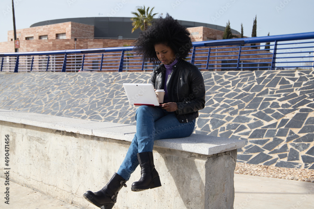 Young and beautiful Afro-American woman with laptop computer shopping online, checking her mail, social networks and working in the park. The woman is wearing casual clothes and drinking coffee, tea.