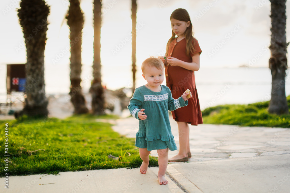 Cute European kid girl in dress on sea. Girl with her sister at sunset on ocean. Walks along seashore in silence at sunset. Kids run stone path near sea, happy childhood and sibling relationships Cute European kid girl in dress on sea. Girl with her sister at sunset on ocean. Walks along seashore in silence at sunset. Kids run stone path near sea, happy childhood and sibling relationships