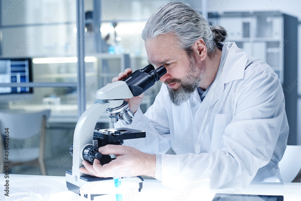 Bearded chemist in lab coat looking through the microscope and examining the virus during scientific experiment at lab Bearded chemist in lab coat looking through the microscope and examining the virus during scientific experiment at lab