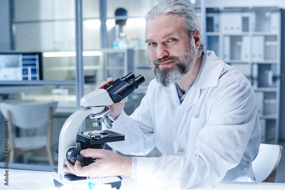 Portrait of bearded medical specialist looking at camera while sitting at table and using microscope in his work at laboratoy Portrait of bearded medical specialist looking at camera while sitting at table and using microscope in his work at laboratoy