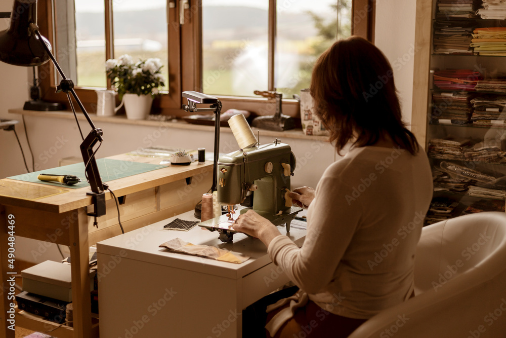 Caucasian brunette woman sewing fabric with a vintage, retro sewing machine. Fashion, creation and tailoring. Process of sewing in atelier or workshop. Special hobby. Cozy interior and warm colors.
