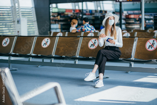 Tourist in mask sitting in airport terminal lounge on empty marked chairs under new normal and covid social distance regulations. Woman sitting in airport lounge and looking at her passport and travel