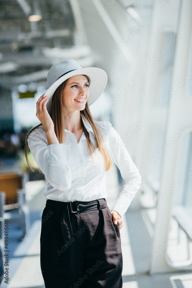 Cheerful woman in stylish blouse and beige pants smiles widely. Attractive blonde girl in white shirt poses with luggage near airport Cheerful woman in stylish blouse and beige pants smiles widely. Attractive blonde girl in white shirt poses with luggage near airport