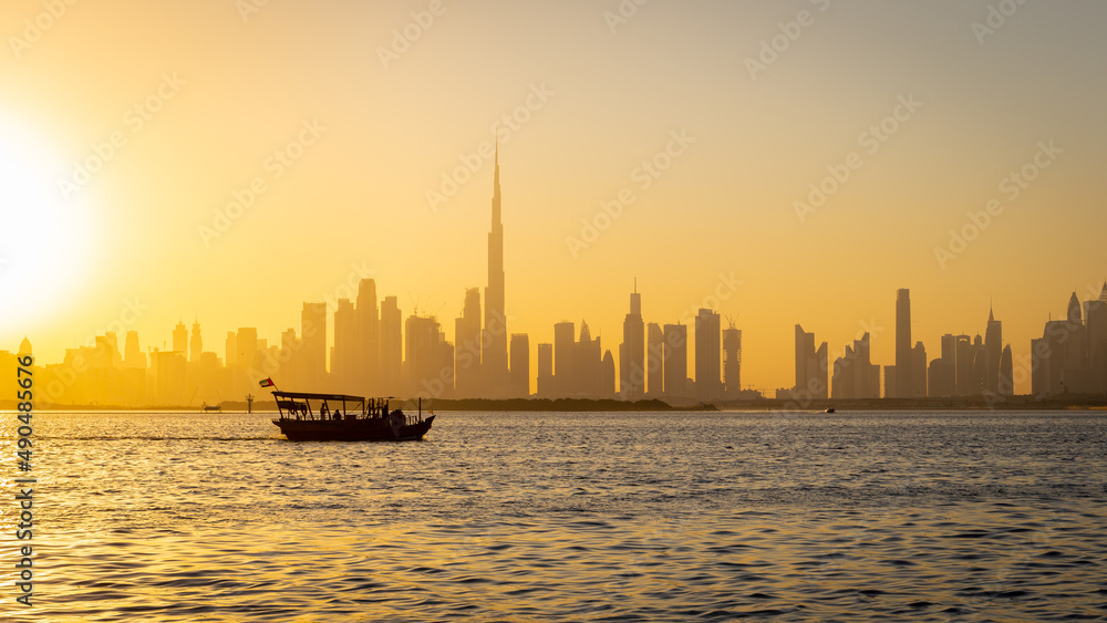 Dubai Downtown skyline panorama during sunset with a silhouette of a tourist boat passing on Dubai Creek Canal, seen from Dubai Creek Harbour promenade.