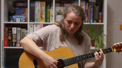 A girl plays an overdub on an acoustic guitar sitting in a room. Behind her is a rack of board games and flowers. A mash-up of music photo