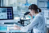 Young woman in lab coat looking through the microscope while sitting at table with computer and test tubes during her work in laboratory