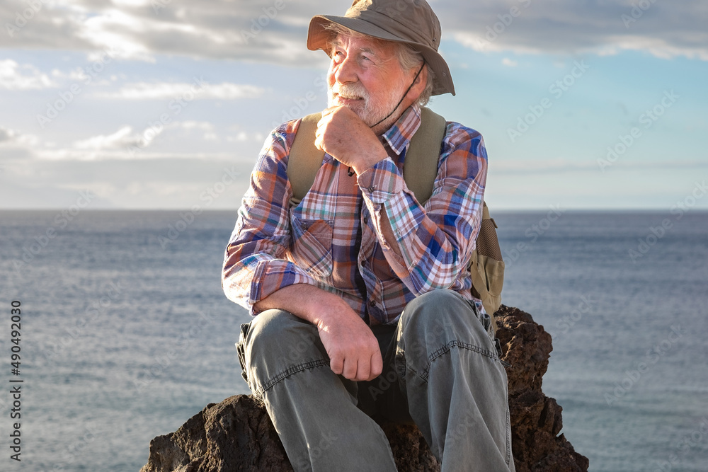 Portrait of senior smiling man resting in outdoor excursion at sea sitting on the cliff looking away. Mature attractive bearded man on a hiking trip enjoying freedom and healthy vacation
