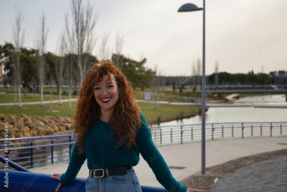 Young woman with red hair, freckles, leaning on a railing and laughing in an outdoor park. Concept happiness, smile, laughter, joy.