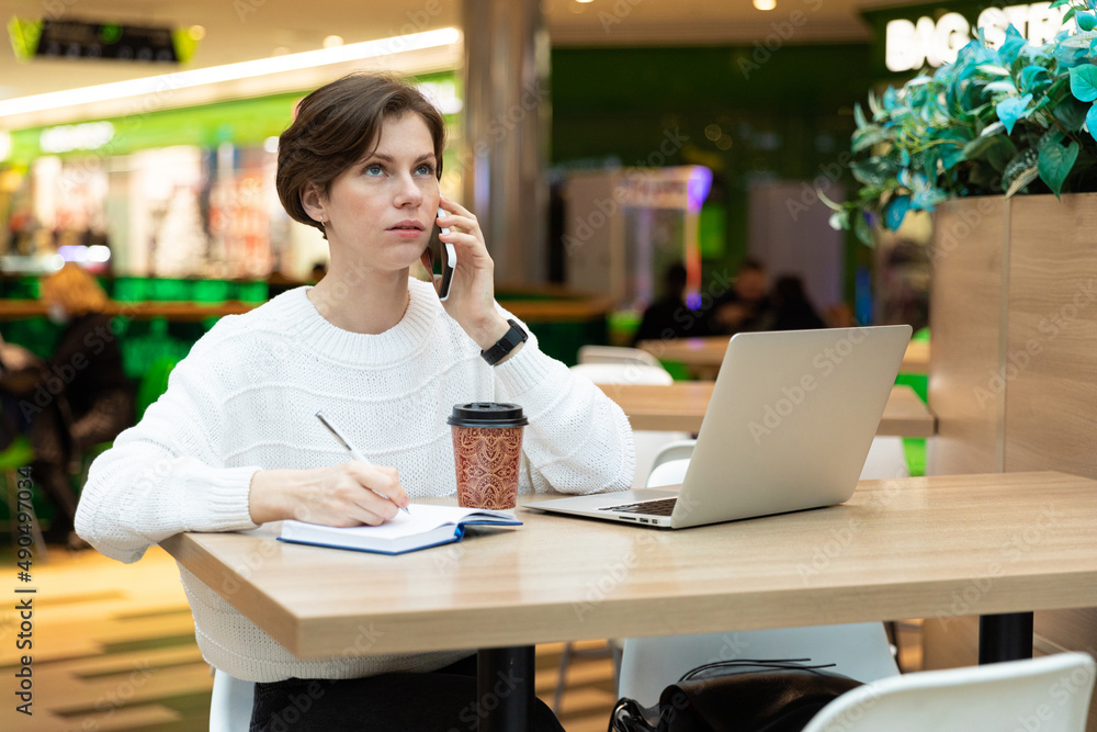Young attractive brunette woman sitting at a table and working at a computer laptop, using mobile phone, writing information at notebook. Freelance and business concept Young attractive brunette woman sitting at a table and working at a computer laptop, using mobile phone, writing information at notebook. Freelance and business concept