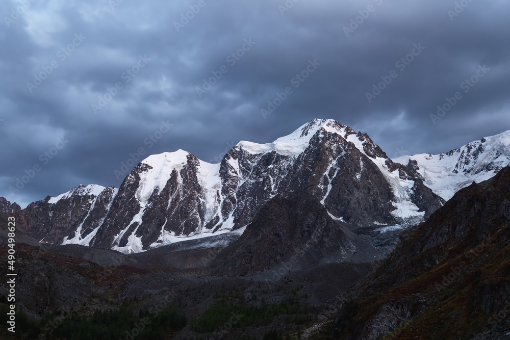 Obraz premium Dark atmospheric alpine landscape with snow-covered mountain top under night sky. Awesome scenery with beautiful pointy peak with snow and high snowy mountain wall with dark low clouds.
