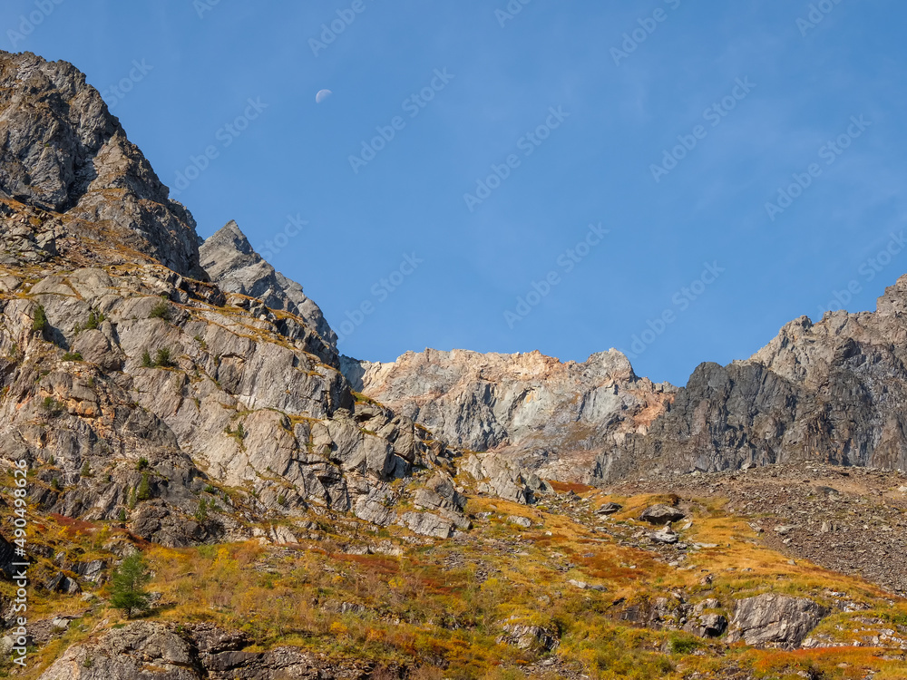 Bright mountain landscape with sharp rocks above yellow slope under blue sky with moon in sunny day. Colorful scenery with gold sunlit sharp rocky mountains. High rocky mountains in golden sunlight.