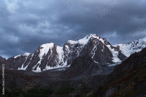 Dark atmospheric alpine landscape with snow-covered mountain top under night sky. Awesome scenery with beautiful pointy peak with snow and high snowy mountain wall with dark low clouds.