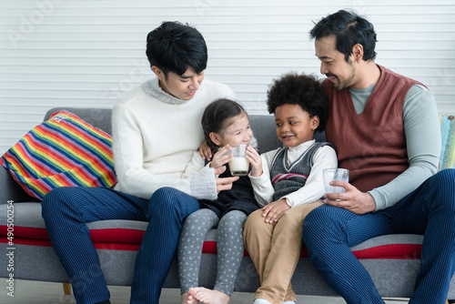 Happy young Asian gay couple with diverse adopted children, African and Caucasian, drinking milk and sitting on sofa at home. Older boy with milk mustache cheer up little girl drink milk. Lgbt family