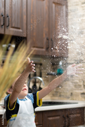 A cute boy of 7 years old in an apron plays with flour at home in the kitchen against the background of kitchen utensils. Selective focus. Portrait