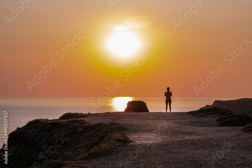 A person stands on the cliff edge of a coast and watches the cloudless sunset.