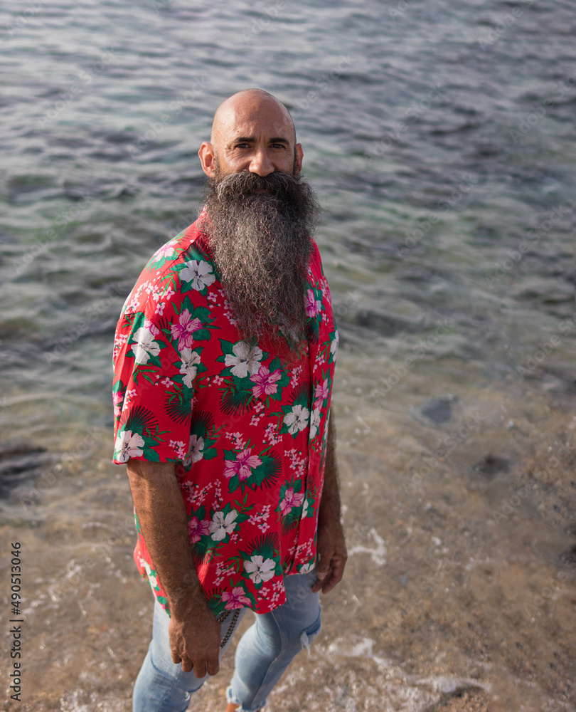 high angle shot of a handsome mature man with long beard and shaved head  dressed hawaiian red shirt standing in a shore of a beach while looking at  the camera Stock Photo |