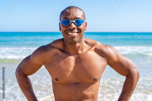 Black swimmer with swimming goggles on and looking at camera smiling by the ocean on a sunny day © Supermelon