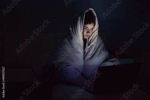 Indoors, a lonely young sad woman sits in the dark on the couch, wrapped herself in a blanket with her head, looks at the laptop screen with emotion of excitement and fear.