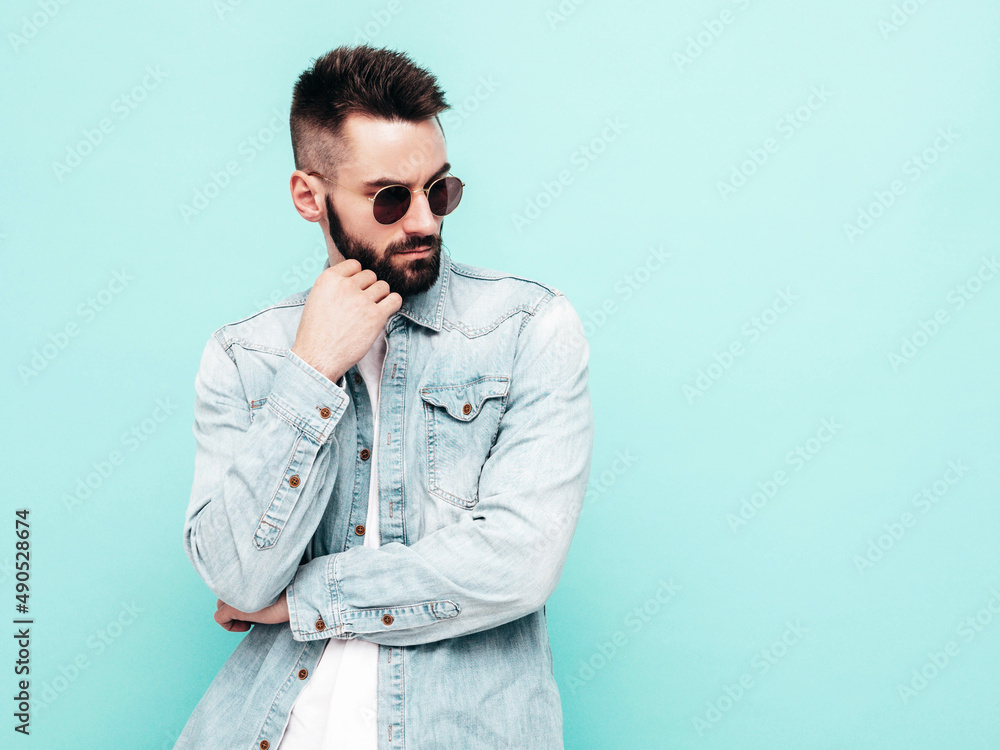 Portrait of handsome confident stylish hipster lambersexual model.Man dressed in jacket and jeans. Fashion male posing near blue wall in studio in sunglasses. Thoughtful Portrait of handsome confident stylish hipster lambersexual model.Man dressed in jacket and jeans. Fashion male posing near blue wall in studio in sunglasses. Thoughtful