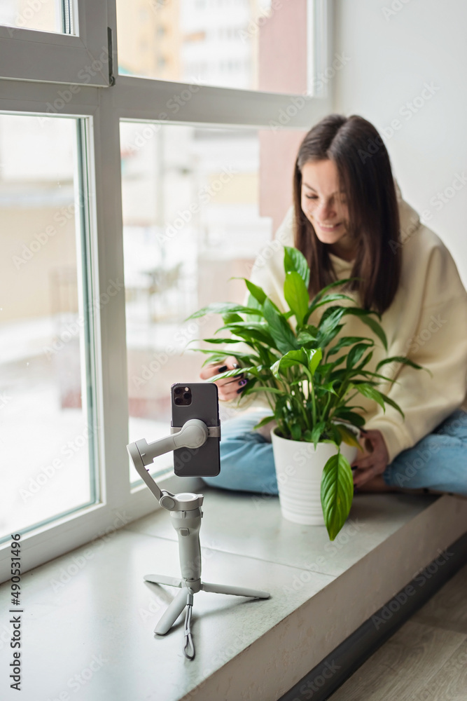 Young beautiful woman influencer blogger blogging about houseplants, filming herself with potted spathiphyllum on a smartphone on a stabilizer tripod. Soft selective focus. Young beautiful woman influencer blogger blogging about houseplants, filming herself with potted spathiphyllum on a smartphone on a stabilizer tripod. Soft selective focus.