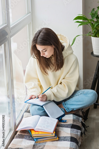 Home schooling. A teenager girl, a student, sits on a windowsill with a bunch of books and writes homework. Distance learning, preparation for exams. Soft selective focus.