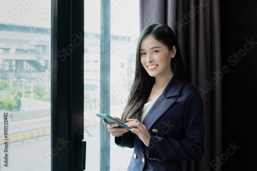 Young businesswoman using smartphone working in hotel room during her business travel. Businesswoman working from hotel room on business trip standing by the window in sunny day. © Avirut S.