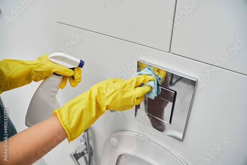 A woman's hand in yellow rubber gloves wipes the toilet flush button with a rag. Sanitary treatment in the bathroom.