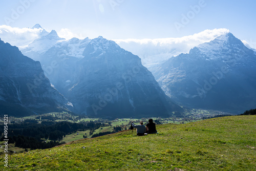 A beautiful Alpine landscape with two tourists sitting on a green meadow hills admiring the distant hilly areas with pine trees below and fantastic mountain ranges covered with clouds on a clear day.