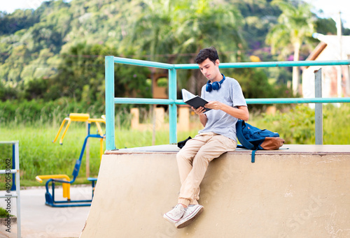 University student sitting and reading a book outdoors, with a backpack by his side.