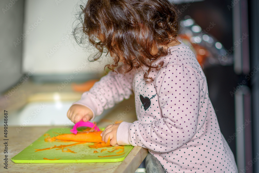 Little cute baby toddler girl in the kitchen peeling carrots with