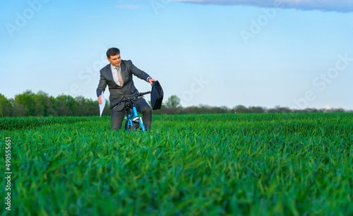 businessman dressed in a business suit, rides a bicycle through a green grass field, he has a briefcase and documents, beautiful nature in spring, business concept