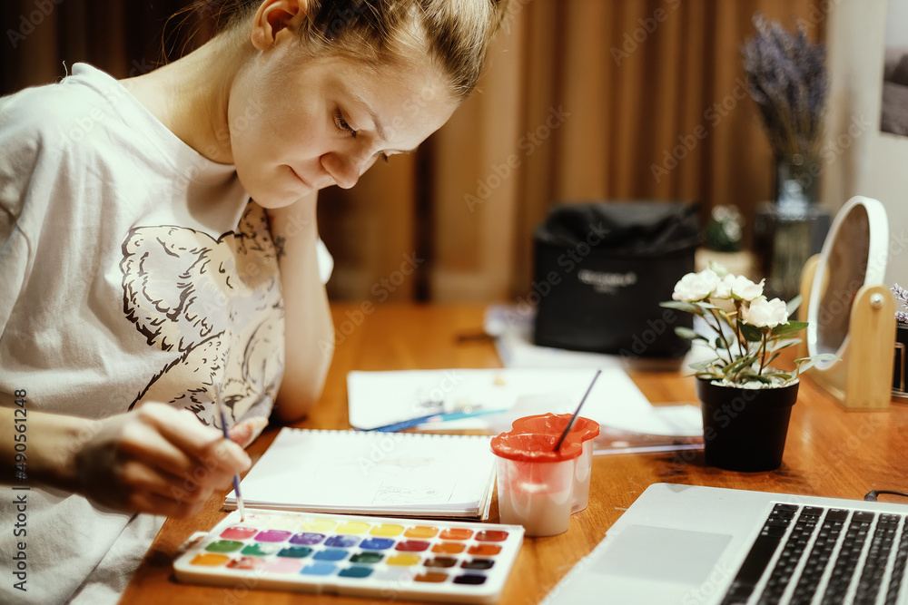 Fototapeta premium Young girl sits at table in her room and paints with watercolor paints. Theme of creativity, inspiration and drawing. Close-up portrait