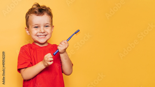 The happy kid is holding a blue toothbrush on a yellow background and smiling showing his teeth. Health care, oral hygiene. A place for your text.