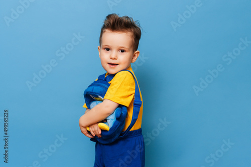 Cute little one cuddles with his backpack on a blue background. Funny kid